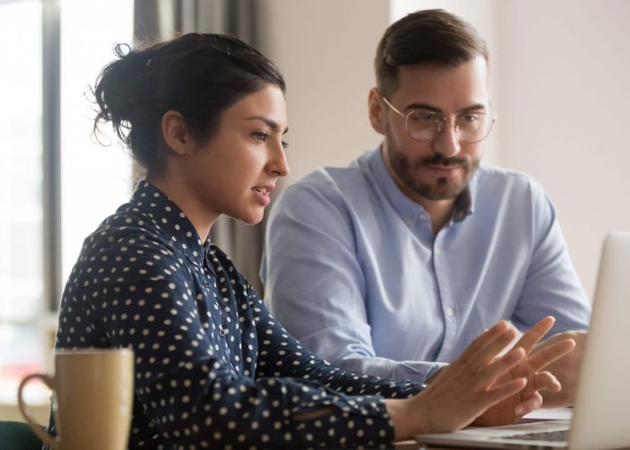 Focused male intern listening to serious indian business woman mentor teacher explaining online strategy looking at laptop computer teach trainee training new worker learning new skill at workplace